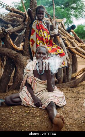 TRIBÙ TOPOSA, SUD SUDAN - 12 MARZO 2020: Fumatrice donna pipa mentre si siede a terra vicino a legname e ragazza in abiti colorati nel villaggio di Toposa Foto Stock