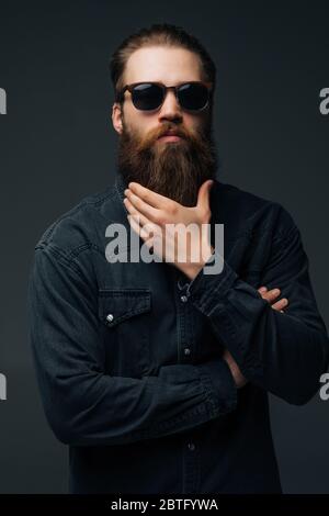 Il giovane uomo in occhiali da sole tocca la sua lunga barba isolata su sfondo grigio Foto Stock