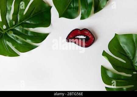 Spilla alla moda a forma di labbra da perle giapponesi, foglie verdi di Monstera su sfondo bianco. Primo piano, disposizione piatta, spazio per la copia. Tendenze concettuali, Foto Stock