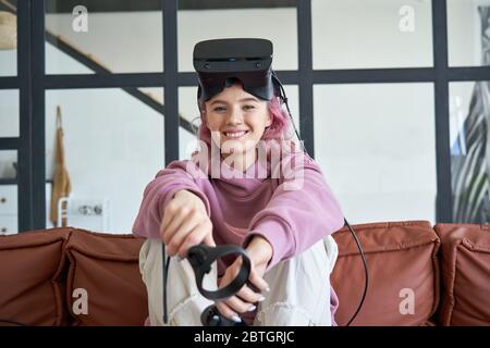 Felice hipster teen girl indossare vr cuffie tenere i controller guardare la telecamera sul divano. Foto Stock