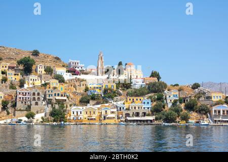 SYMI, Grecia - 27 giugno 2019: Vista della costa dell'isola di Symi con la città con case multicolore, alberi e popoli e baia con le barche in estate giorno Foto Stock