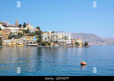 SYMI, Grecia - 27 giugno 2019: Vista della costa dell'isola di Symi con la città con case multicolore, alberi e popoli e baia con le barche in estate giorno Foto Stock