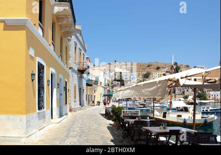 SYMI, Grecia - 27 giugno 2019: Vista della costa dell'isola di Symi con la città con case multicolore, alberi e popoli e baia con le barche in estate giorno Foto Stock