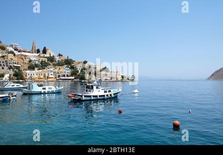 SYMI, Grecia - 27 giugno 2019: Vista della costa dell'isola di Symi con la città con case multicolore, alberi e popoli e baia con le barche in estate giorno Foto Stock