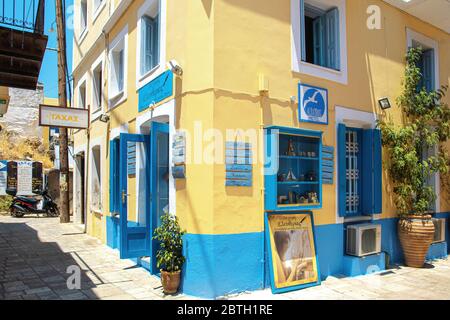 SYMI, Grecia - 27 giugno 2019: Via della città di Symi nell'isola della Grecia con le finestre blu e le porte di caffè, hotel e negozio e fiori pentole Foto Stock