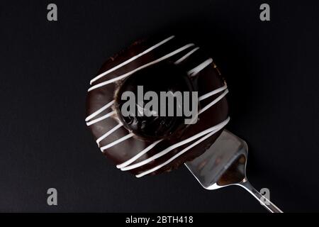 Una ciambella di cioccolato con glassa bianca a strisce su sfondo nero. Una fetta di torta d'argento si trova sotto la ciambella Foto Stock