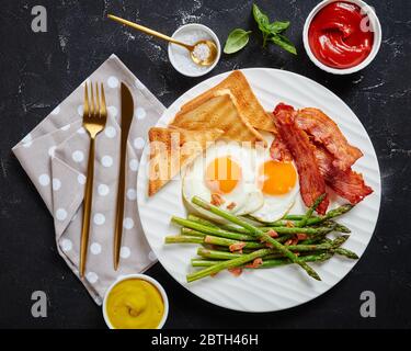 uova fritte, pancetta croccante, toast, asparagi su un piatto bianco con forchetta dorata e coltello su un tavolo di cemento con ketchup, senape, vista orizzontale da Foto Stock