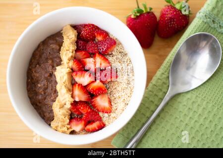 Farinata d'avena al cacao al cioccolato con farina di mandorle, fragole tritate e burro di arachidi cremoso su una superficie di legno. Fragola, mandorle, tovagliolo e cucchiaio Foto Stock
