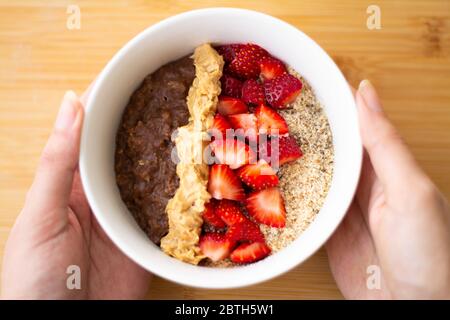 Farinata d'avena al cacao al cioccolato con farina di mandorle, fragole tritate e burro di arachidi cremoso su una superficie di legno. Fragola, mandorle, tovagliolo e cucchiaio Foto Stock