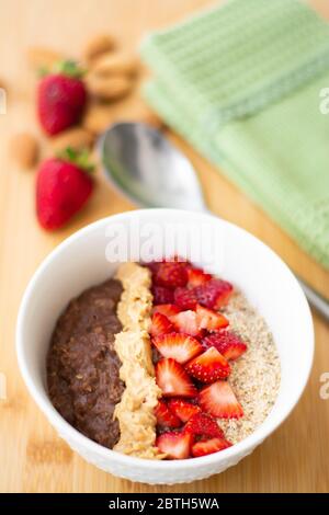 Farinata d'avena al cacao al cioccolato con farina di mandorle, fragole tritate e burro di arachidi cremoso su una superficie di legno. Fragola, mandorle, tovagliolo e cucchiaio Foto Stock