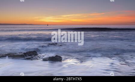 Onde che rotola sopra le rocce sulla costa, mentre il sole tramonta sull'orizzonte Foto Stock