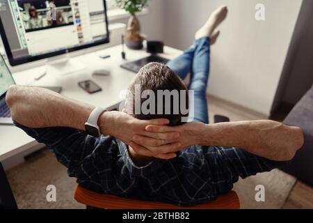 Uomo che si rilassa mentre lavora a distanza da casa tenendo le gambe sul tavolo guardando il computer che allunga le mani dietro la testa Foto Stock