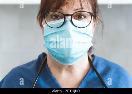 Primo piano ritratto di una donna seria medico con maschera protettiva e uniforme medica - fuoco sugli occhi con sfondo grigio Foto Stock