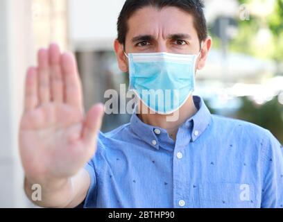 FERMARE COVID-19 uomo che indossa la maschera chirurgica sul viso facendo GESTO DI ARRESTO guardando la fotocamera all'esterno. Giovane uomo con maschera che mostra palmo aperto della mano al Foto Stock