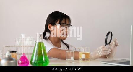 Foto di giovane adorabile ragazza della scuola che tiene una lente d'ingrandimento mentre fa un esperimento scientifico e seduto al tavolo bianco moderno con la bianca Foto Stock