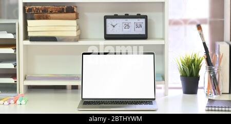 Foto di computer portatile con schermo bianco vuoto che si mette sulla scrivania bianca studente e circondato da orologio, libri, pianta in vaso, matite in vaso di vetro, no Foto Stock