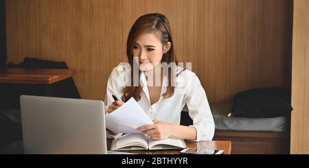 Foto di giovane donna elegante indossata su camicia bianca, incentrata sul suo lavoro mentre si siede di fronte al suo computer portatile alla scrivania in legno sopra con Foto Stock