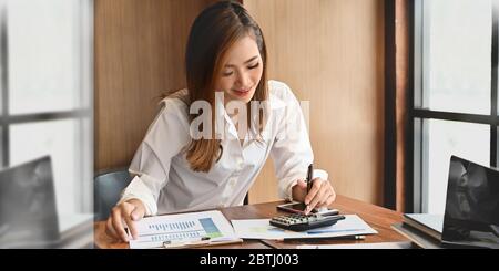 Foto di giovane donna elegante indossata su camicia bianca, incentrata sul suo lavoro mentre si siede di fronte al suo computer portatile alla scrivania in legno sopra con Foto Stock