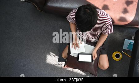 Immagine dall'alto di un uomo creativo che lavora con un tablet bianco con computer a schermo vuoto che si mette sui giri mentre prende appunti e si siede sul sedile Foto Stock