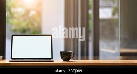 Foto del computer portatile con schermo bianco vuoto e tazza di caffè che si mette su bancone bar in legno sopra una comoda parete di vetro del soggiorno come sfondo. Foto Stock