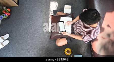 Immagine dall'alto dell'uomo creativo che si disegna su un tablet con schermo bianco e penna stilo mentre si siede al pavimento del salotto che è circondato da Foto Stock