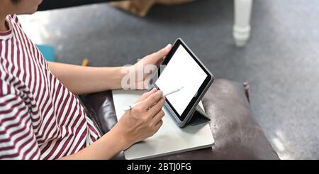 Immagine ritagliata di un uomo creativo che si sta disegnando sul tablet del computer con una penna stilo mentre si siede sul divano in pelle con un tranquillo salotto come sfondo. Foto Stock