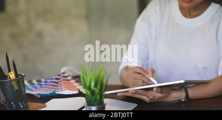 Immagine ritagliata di una donna creativa in una maglietta bianca che tiene e disegna il tablet del computer utilizzando una penna stilo mentre si siede alla scrivania di legno Foto Stock