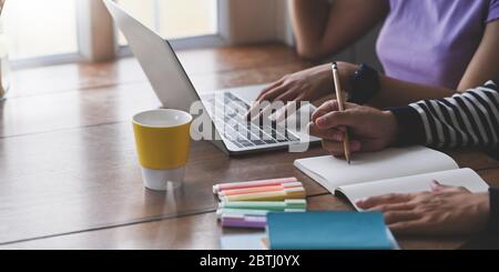 Immagine ritagliata di una giovane coppia che fa i compiti insieme mentre si siede di fronte al computer portatile che si mette su scrivania in legno e surround Foto Stock