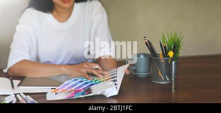 Foto di un giovane designer grafico che tiene una penna stilo mentre seleziona il colore dalla guida colori e seduto davanti al tablet del computer con schermo vuoto Foto Stock