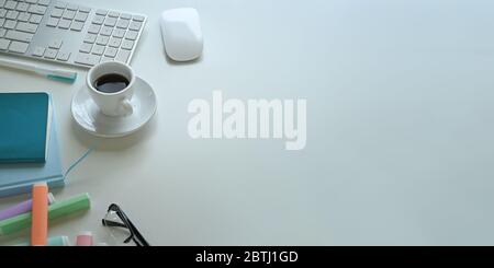 Immagine dall'alto della tazza da caffè che si trova sulla scrivania bianca circondata da mouse wireless, tastiera, pennarelli, occhiali, notebook, note e p Foto Stock