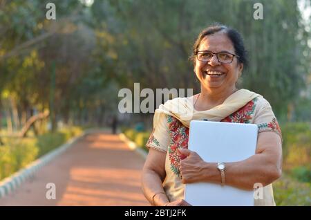 Ritratto di una donna indiana senior sicura in pensione che porta un computer portatile in un parco con il kamiz bianco della guerra di salamaglia. Concetto - alfabetizzazione digitale in India fo Foto Stock