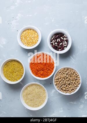 Riso, bulgur, fagioli, piselli, lenticchie e ceci in ciotole bianche su sfondo grigio. I cereali sono una fonte di energia. Concetto di cibo sano. Vista dall'alto. Foto Stock