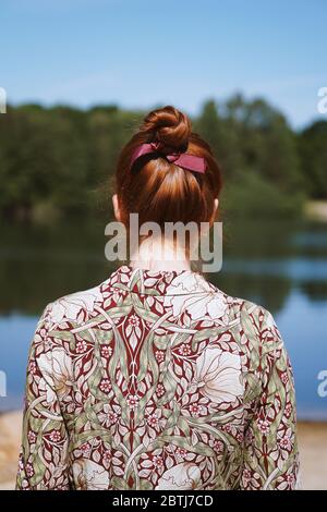 vista posteriore di giovane donna anonima con abito floreale e capelli rossi in piedi vicino al lago in solitudine o depressione Foto Stock