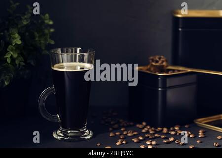 Tazza trasparente di caffè nero caldo sul tavolo nero. Chicchi di caffè in una lattina nera con un cucchiaio per misurare l'oro. Foto Stock