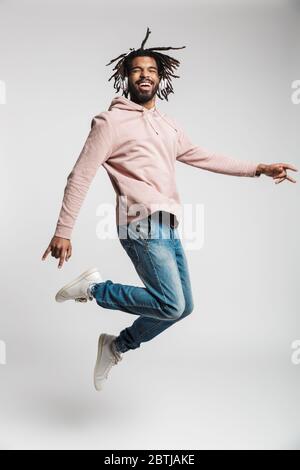Ritratto a tutta lunghezza di un giovane africano allegro che indossa una felpa con cappuccio che salta isolato su sfondo bianco, ascoltando musica con le cuffie Foto Stock