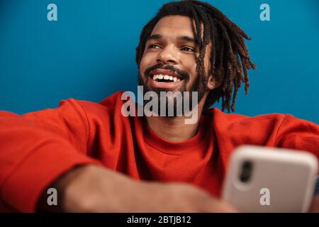 Primo piano ritratto di un giovane africano sorridente che indossa un pullover seduto isolato su sfondo blu, utilizzando il telefono cellulare Foto Stock