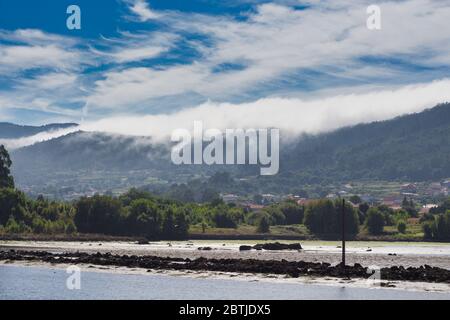 Un mare di nuvole bianche che scende lungo le montagne fino al villaggio di Noia in Galizia, Spagna e raggiunge il mare (bassa marea). Foto Stock