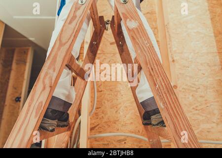 Lavoratore appaltatore di miglioramento domestico che rimane sulla scala di legno. Attrezzature di supporto per cantieri edili. Tema industriale. Foto Stock