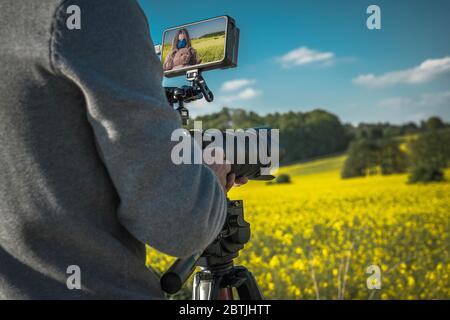 Realizzazione di film per esterni con fotocamera digitale. Video fotografo che scatta foto di ragazza caucasica con scenico paesaggio colorato in background. Digitale moderno Foto Stock