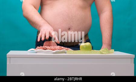 l'uomo con una pancia grassa sceglie tra salsiccia e mela verde. Il concetto di dieta e di volontà. Foto Stock