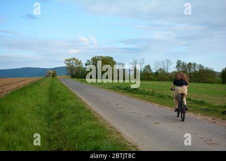 Giovane donna da dietro, si siede su una bicicletta e guida su una strada solitaria in natura verde con cielo blu Foto Stock