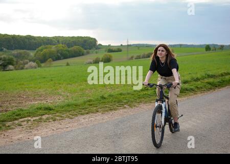 Una giovane donna si siede su una bicicletta e guida su una strada solitaria in natura verde con cielo blu Foto Stock