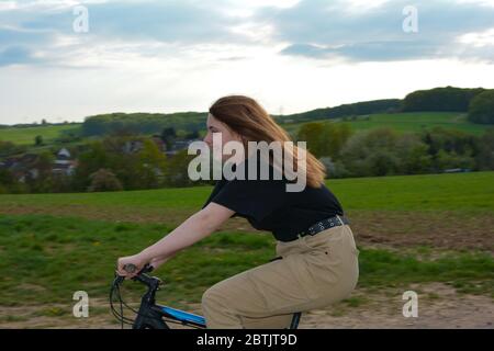 Giovane donna dal lato, si siede su una bicicletta e guida su una strada solitaria di campagna in natura verde con cielo blu e un villaggio sullo sfondo Foto Stock