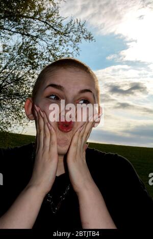 Giovane donna con capelli molto corti e unghie lunghe, mani sulle guance, fa un viso divertente e si leva in natura Foto Stock