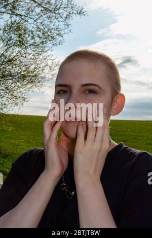 Giovane donna con capelli molto corti e unghie lunghe, mani sul viso serio, si erge in natura Foto Stock