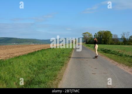 La giovane ragazza cavalcava uno skateboard su una strada di campagna in natura con spazio di copia e cielo blu Foto Stock