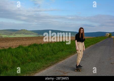 La giovane ragazza cavalcava uno skateboard su una strada di campagna in natura verde, con cielo blu e molti spazi per le copie Foto Stock