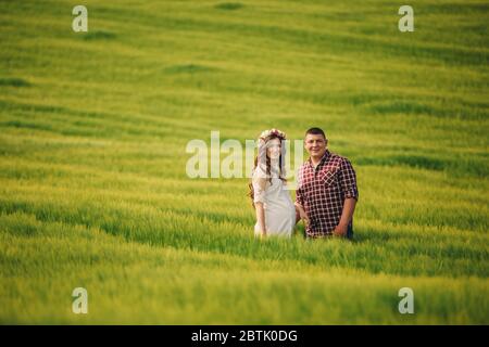 attesa bambino. Genitorialità. La giovane donna incinta e il marito sono felici di tenere le mani, andando all'aperto nel campo di sfondo verde erba. S Foto Stock