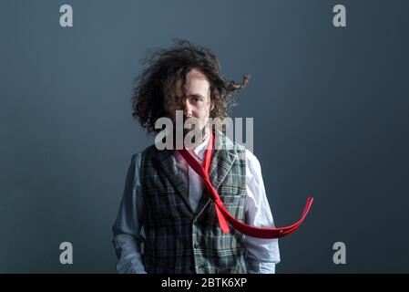 studio ritratto in movimento di un giovane ragazzo bearded, curvly haired di trent'anni, in un gilet plaid, e un cravatta rosso volante, ondeggiare i capelli lunghi. Su un bac grigio Foto Stock