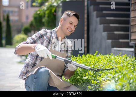 Felice maschio dai capelli scuri in guanti protettivi potando cespugli fuori, accovacciando Foto Stock